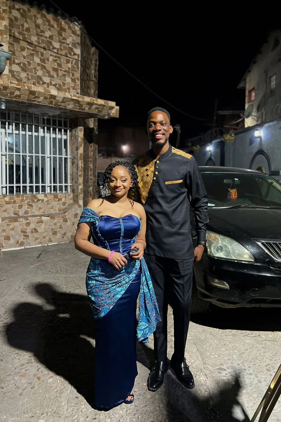 Divine en robe bleue et pagne, Matthieu en tenue noire et dorée, côte à côte dans une rue de nuit